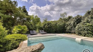 Une piscine entourée de chaises longues, de verdure luxuriante et d’un grand rocher décoratif.
