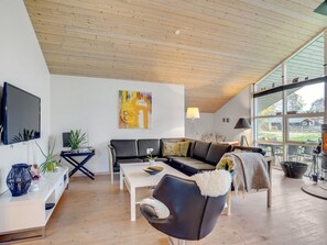 A modern living room with a wooden ceiling, a black leather sofa, a white coffee table, and a television on the wall.