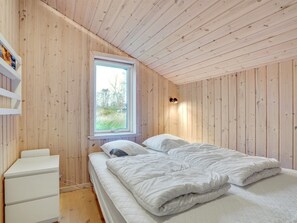 A bedroom with a wooden ceiling, a window with a view of greenery, a bed with white bedding, and a white bedside table.