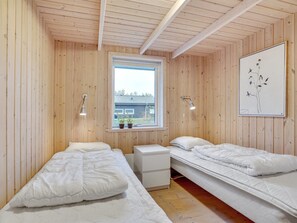 A room with wooden walls and a sloped ceiling, featuring two beds with white bedding, a small bedside table, and a window with a view of a building outside. - "Elni" - 900m from the sea by Interhome (Hals)