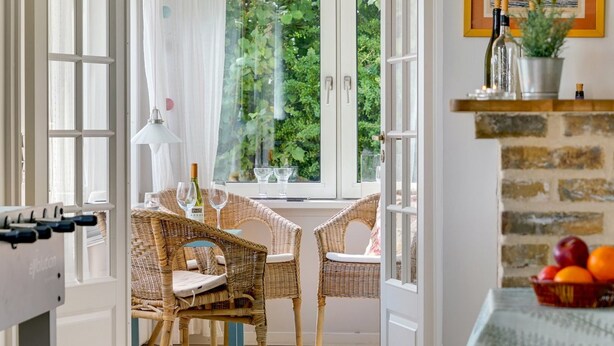 Une pièce avec des fauteuils en osier, une table avec des verres à vin et une vue sur la verdure grâce à de grandes fenêtres.