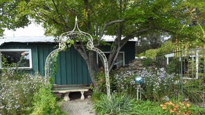 Un sendero en un jardín que lleva a un banco bajo un arco decorativo, rodeado de plantas y un muro de piedra.