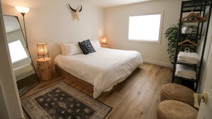 Un dormitorio con cama, dos lámparas de noche, un espejo, una alfombra, suelo de madera, un estante con toallas y una planta.