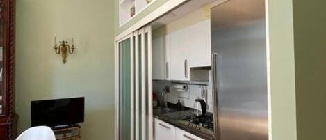 A compact kitchen with white cabinets, a stainless steel refrigerator, and a dining area with a wooden table and white chairs.