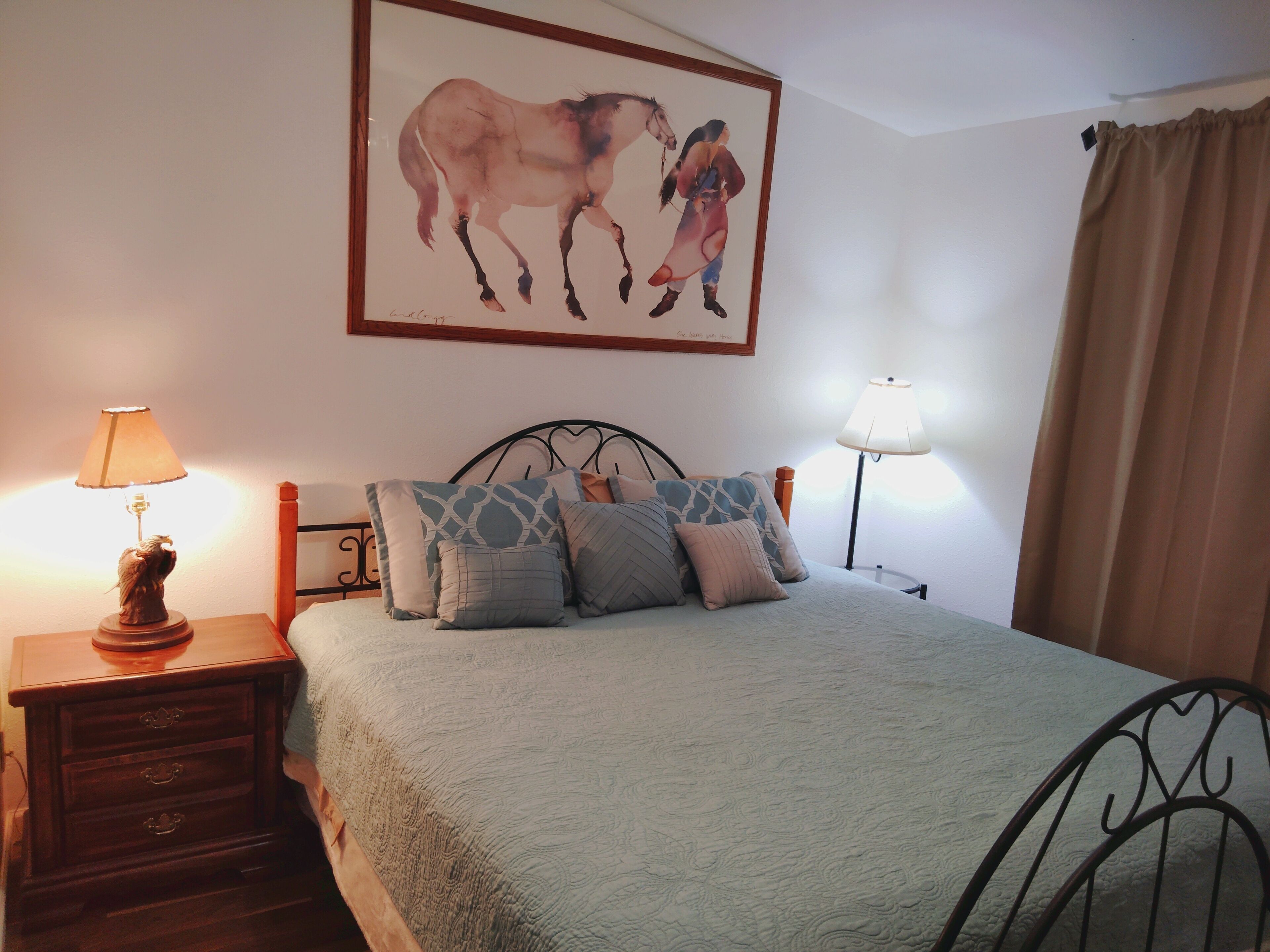 A bedroom with a bed, two bedside lamps, a nightstand, a framed picture on the wall, and a window with curtains.