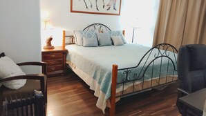 Une chambre à coucher avec un lit, un plancher en bois, une table de chevet, une lampe, une chaise, un bureau et un tableau au mur.