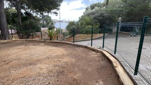 A fenced outdoor area with a dirt ground, surrounded by trees and a view of a body of water. - RENECROS 1 à 30 m de la mer Very Large Terrace Wifi Clim Parking (BANDOL)