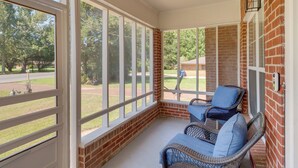 A covered porch with wicker furniture and a view of a brick building and greenery. - Renovated Louisiana Rental Near Mississippi River (Ferriday)