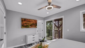 A modern bedroom with a large bed, a ceiling fan, a painting, and a view of the outdoors. - Tree-Lined Lake Arrowhead Cabin ~ 1 Mi to Village! (Lake Arrowhead)