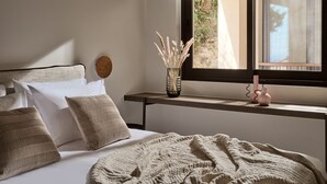 A bedroom with a bed, pillows, a bedside table with a vase of dried plants, and a window with a view of greenery.