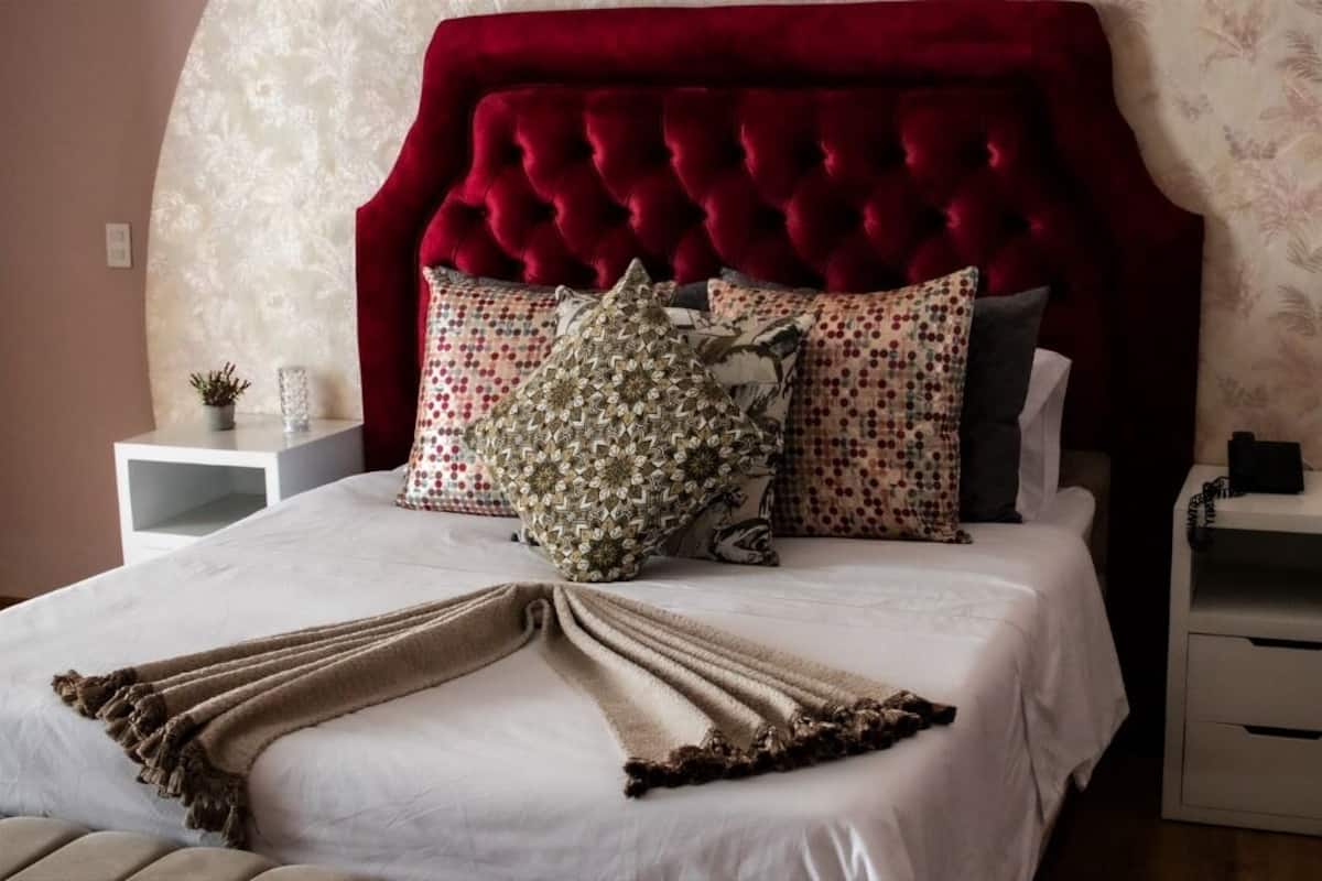 Une chambre à coucher avec une tête de lit rouge à motifs capitonnés, un couvre-lit blanc, plusieurs coussins à motifs et deux tables de chevet avec des tiroirs.