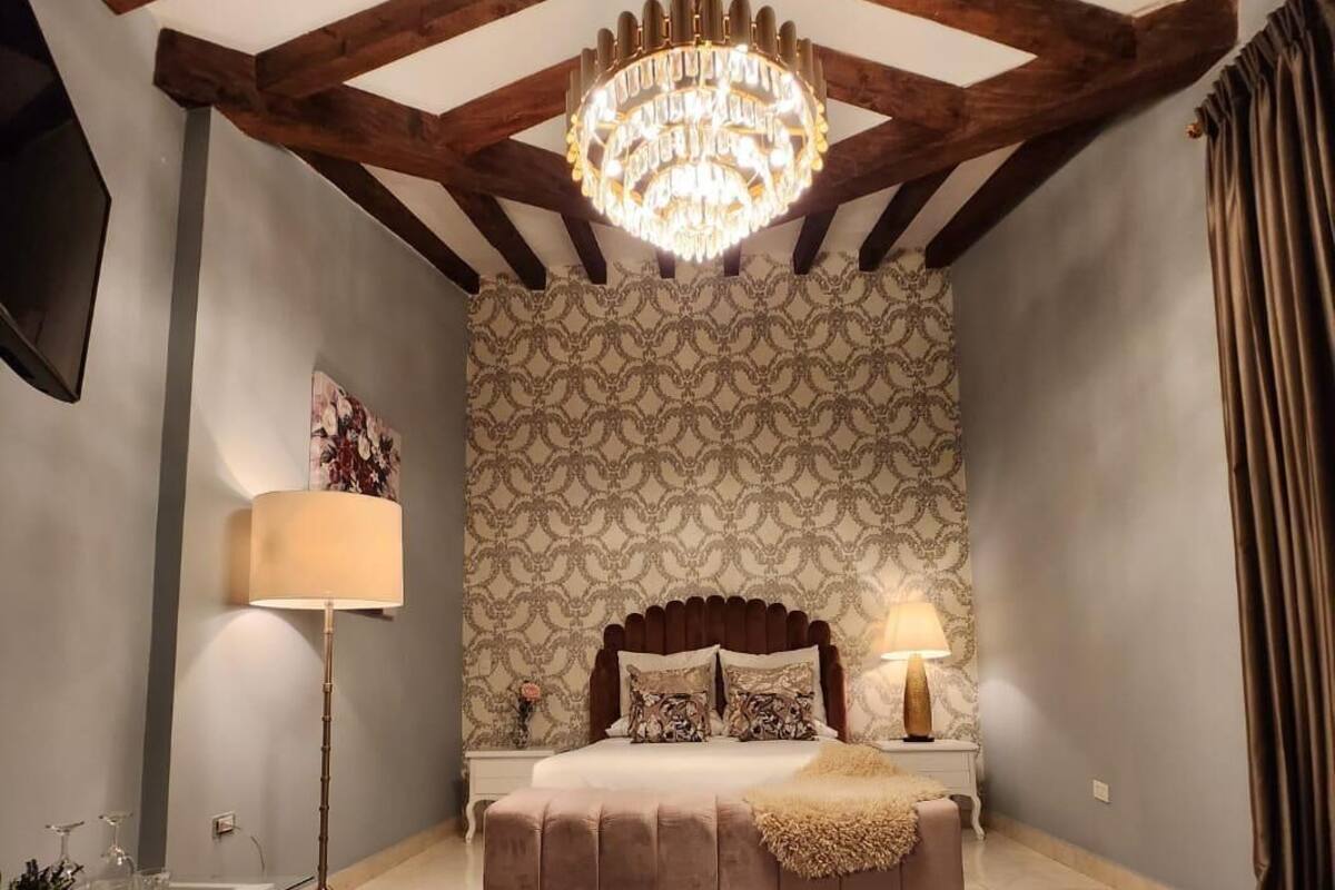 Une chambre à coucher avec un grand lit, un lustre, un papier peint à motifs et un tapis rouge foncé.