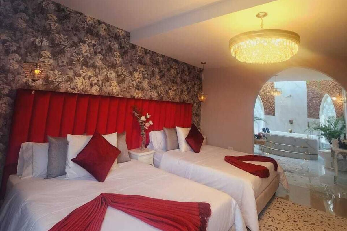 Une chambre d’hôtel avec deux lits, une tête de lit rouge, un lustre et une vue sur l’espace piscine.
