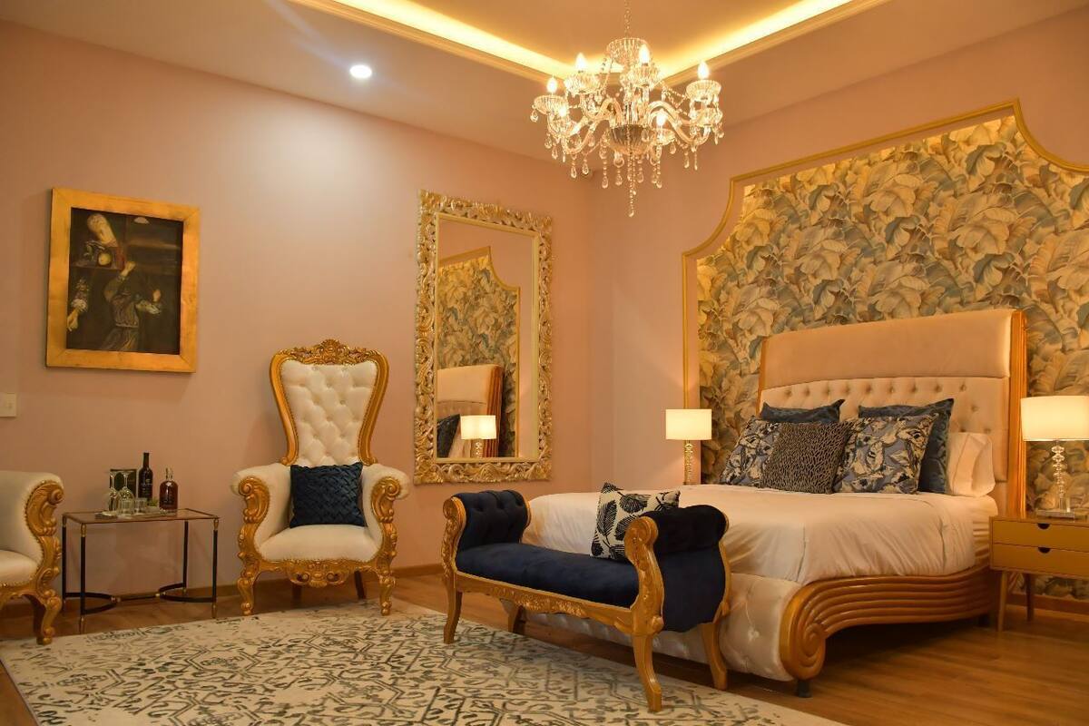 Une chambre à coucher avec un grand lit, deux fauteuils, une chaise longue, un lustre et un papier peint à motifs floraux.
