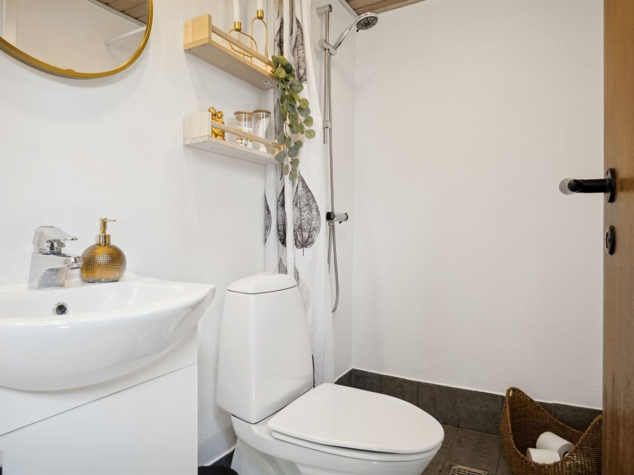 Une salle de bain avec un lavabo, des toilettes et une douche, le tout de couleur blanche. Au-dessus du lavabo, on trouve des étagères où sont rangés les produits de toilette, ainsi qu’un miroir avec un cadre doré.