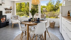 Una cocina moderna con armarios blancos, un comedor con una mesa redonda de madera y sillas blancas, una estufa de leña empotrada y ventanas grandes que ofrecen vistas al exterior.