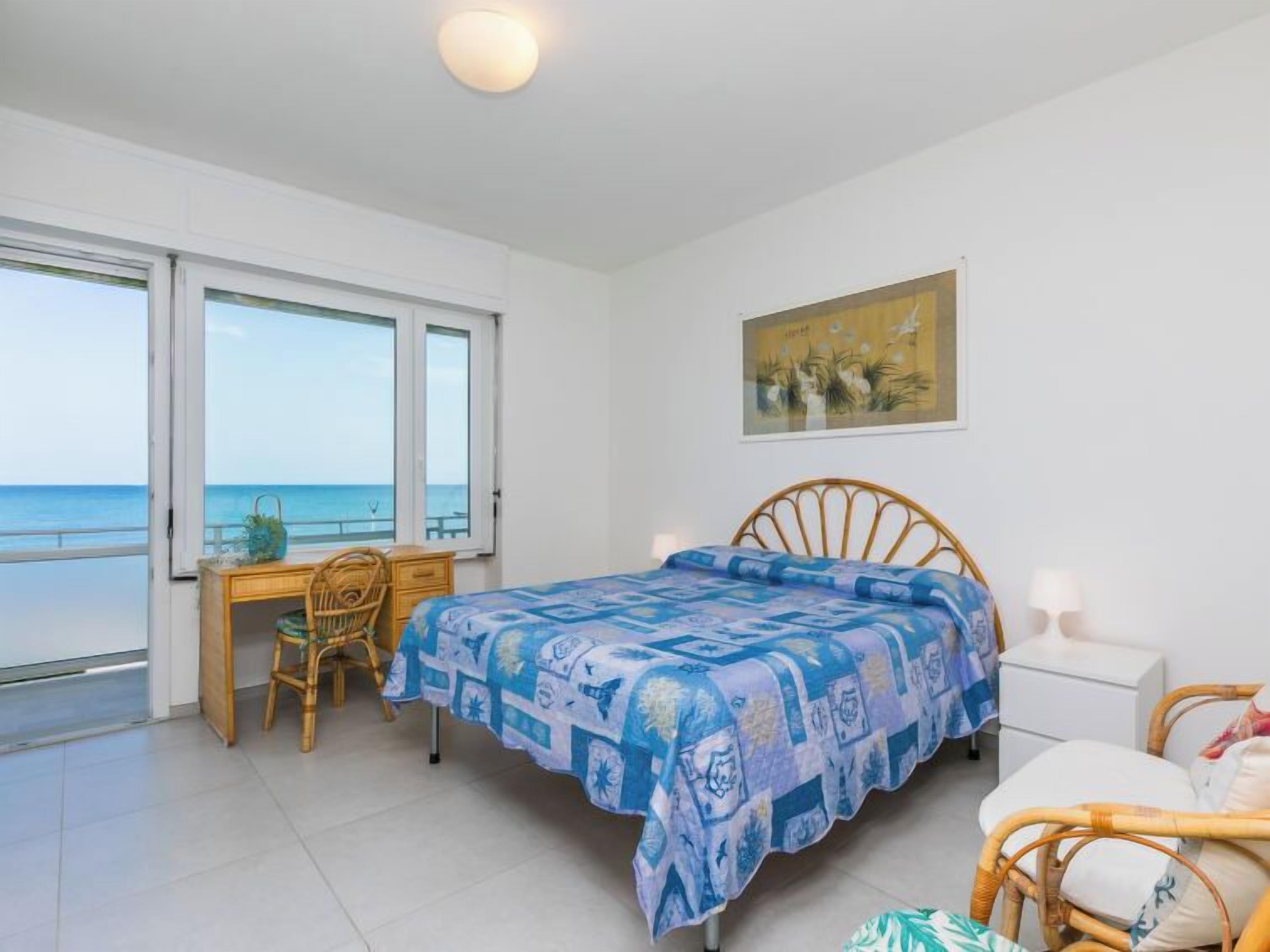 Un dormitori amb un llit, un escriptori i una cadira. Hi ha una finestra gran amb vista al mar.