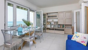 A modern living room with a glass dining table, metal chairs, a blue sofa, and a bookshelf. - Massimo Beach House by Interhome (Castiglioncello)