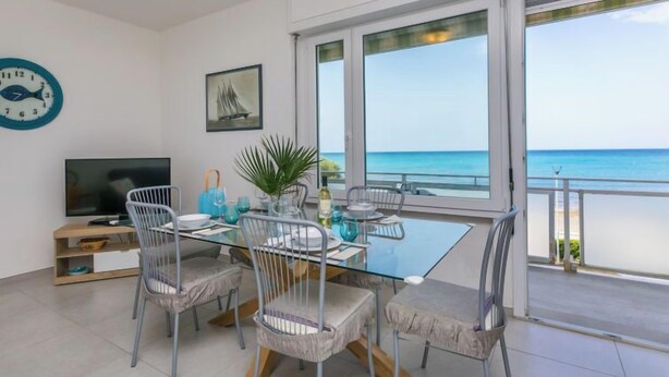 A modern dining area with a glass table, metal chairs, and a view of the sea through large windows. - Massimo Beach House by Interhome (Castiglioncello)