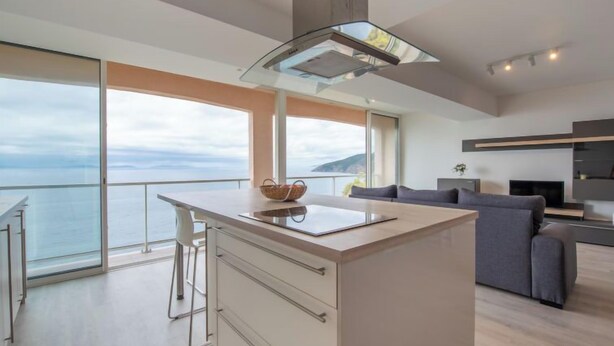 A modern kitchen with a large island, a dining area, and a sofa. There is a view of the sea through large windows. - Le Grand Large by Interhome (Cavalaire)