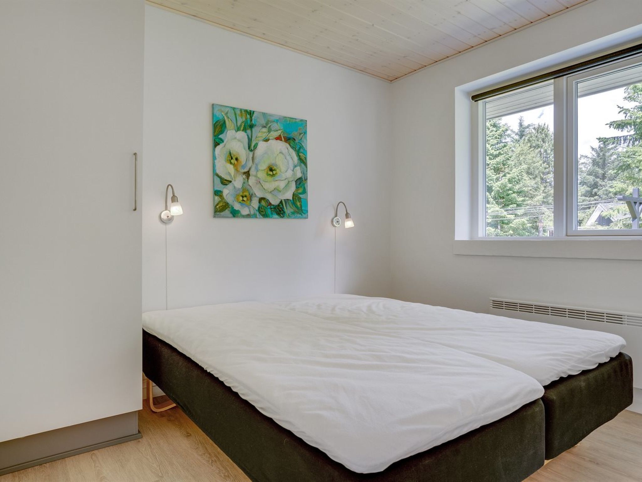 A modern bedroom with a large bed, two wall-mounted lights, a window with a view of trees, and a painting of flowers on the wall.