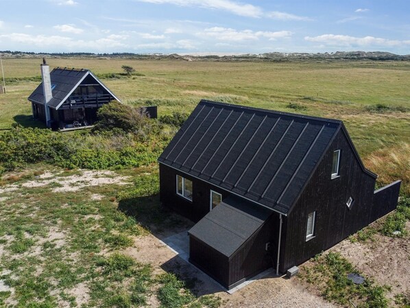 A modern, dark-colored house with a sloped roof, situated in a grassy field with a distant view of dunes.