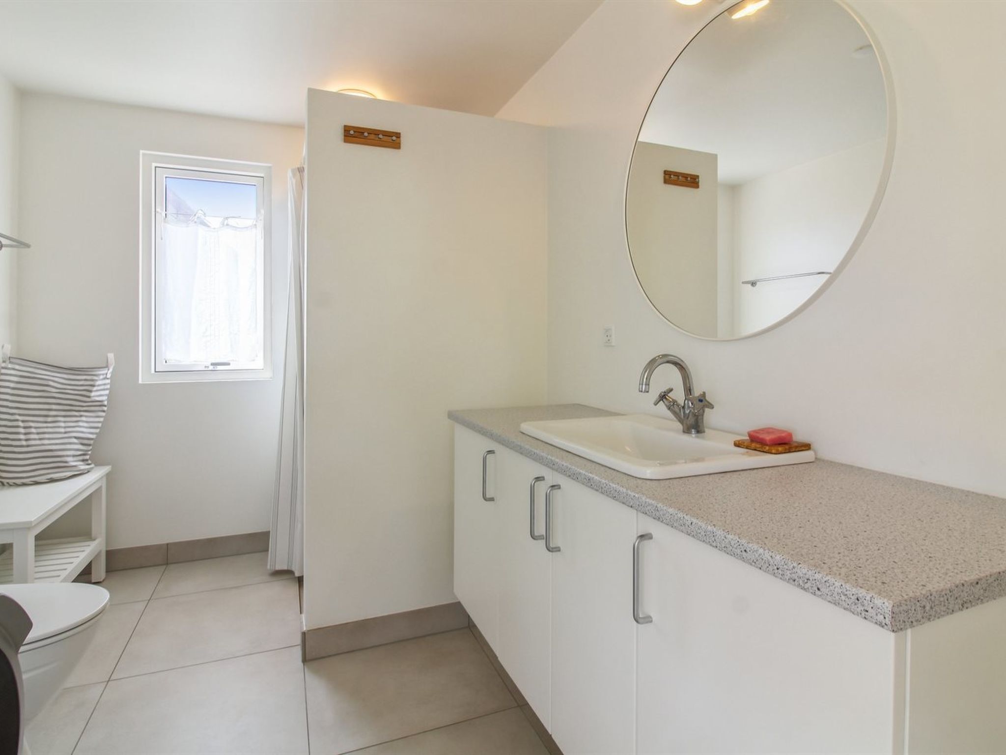 A modern bathroom with a white sink, a round mirror, and a window with curtains.