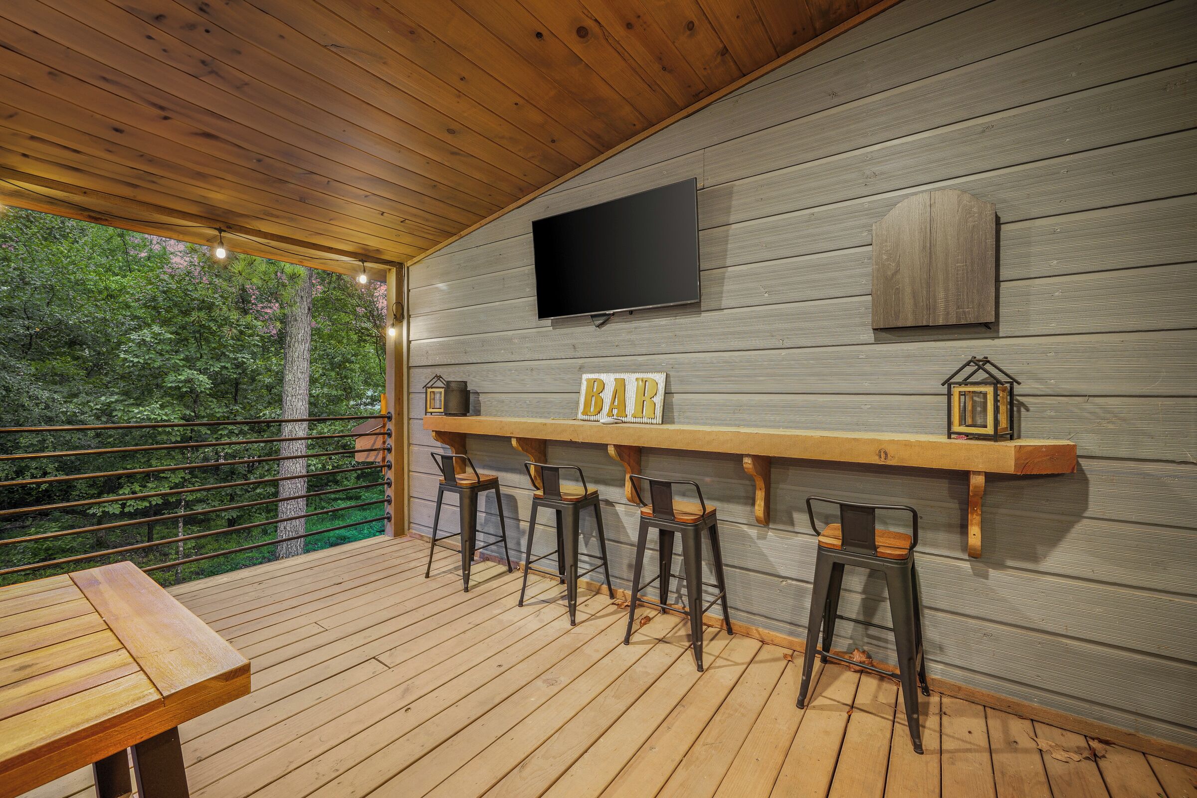 A wooden deck with a bar area, bar stools, a TV mounted on the wall, and a wooden table.