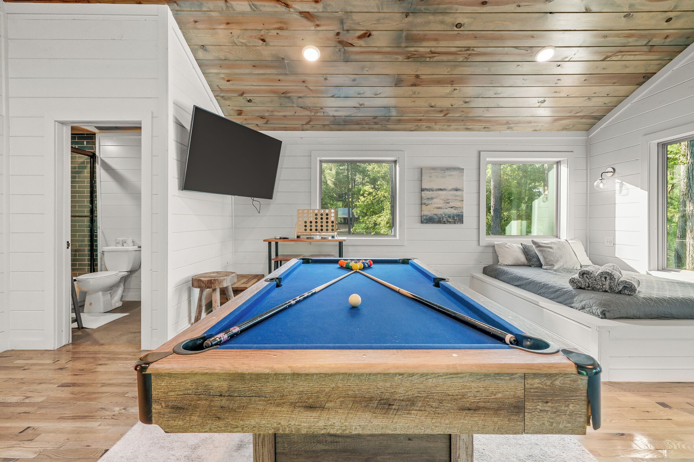 A pool table in a room with white walls, wooden ceiling, and large windows.