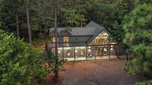 A two-story house surrounded by dense forest, with a gravel driveway and multiple windows illuminated at dusk.