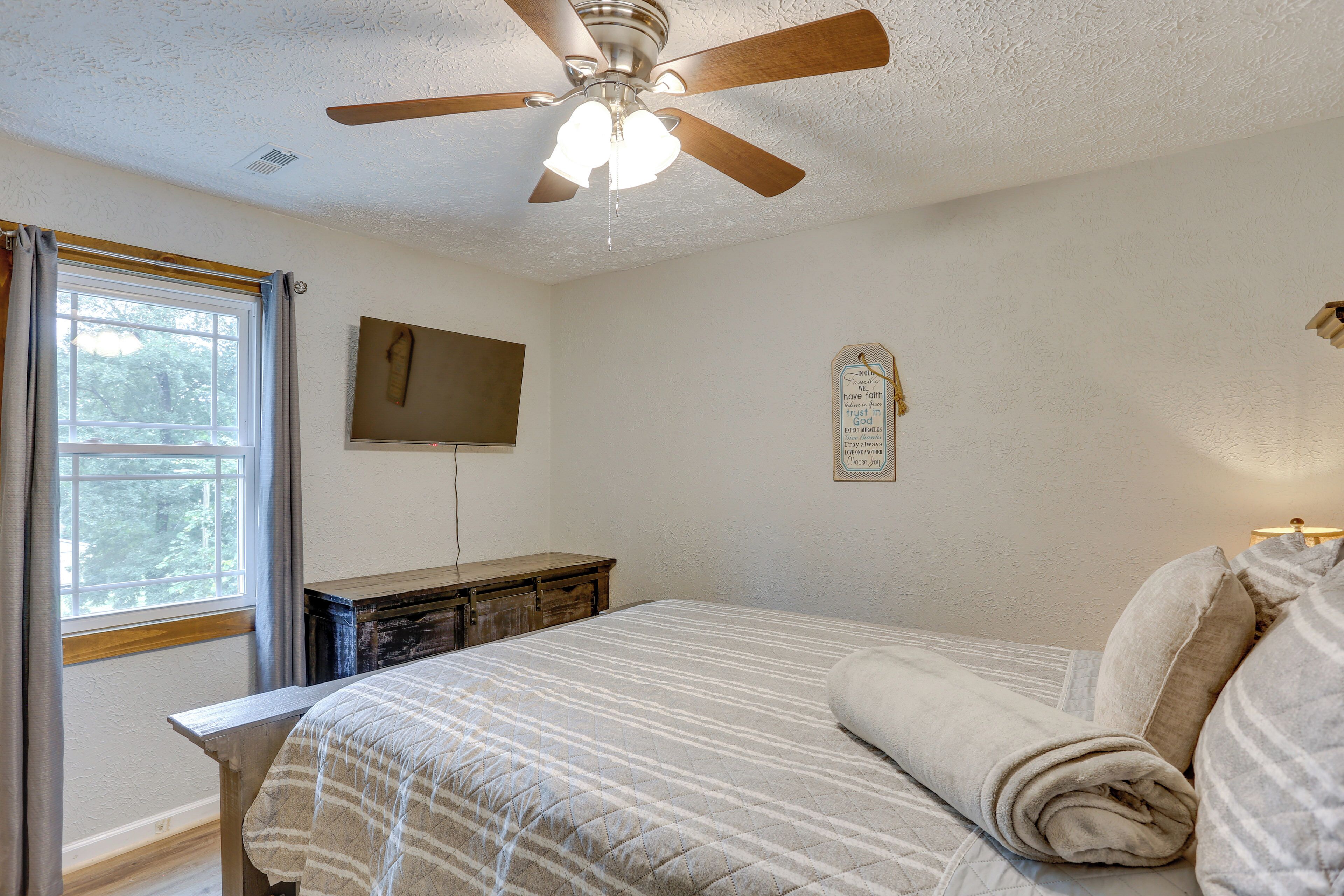 A bedroom with a bed, a ceiling fan, a window with curtains, a wooden nightstand, and a wall-mounted TV.