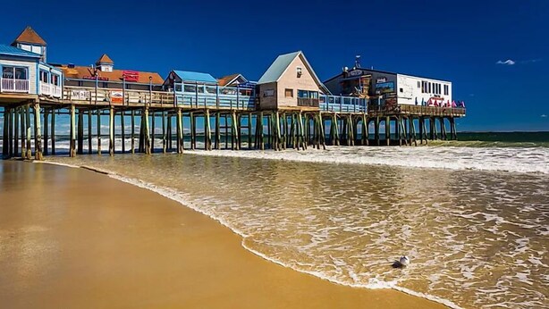A pier with buildings extending over the water, a sandy beach, and a clear blue sky.