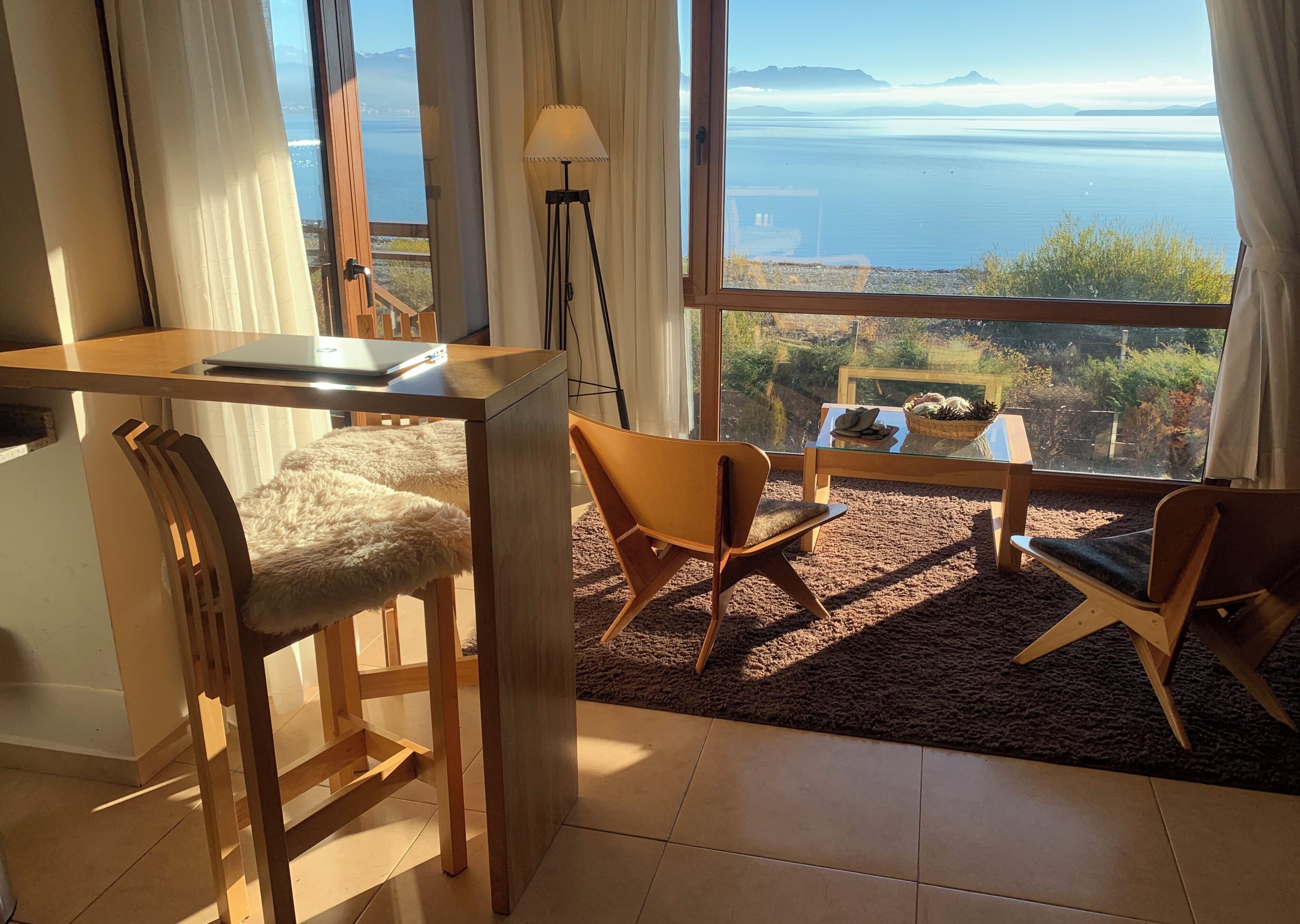 A room with a view of the sea, a desk with a laptop, two chairs, a lamp, and a small table with plants.