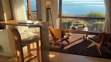 A room with a view of the sea, a desk with a laptop, two chairs, a lamp, and a small table with plants.