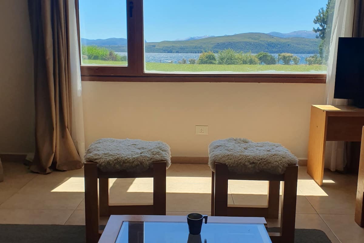 Una habitación con un gran ventanal, dos taburetes de madera con cojines mullidos, una mesa con superficie de vidrio y un televisor sobre un mueble de madera.