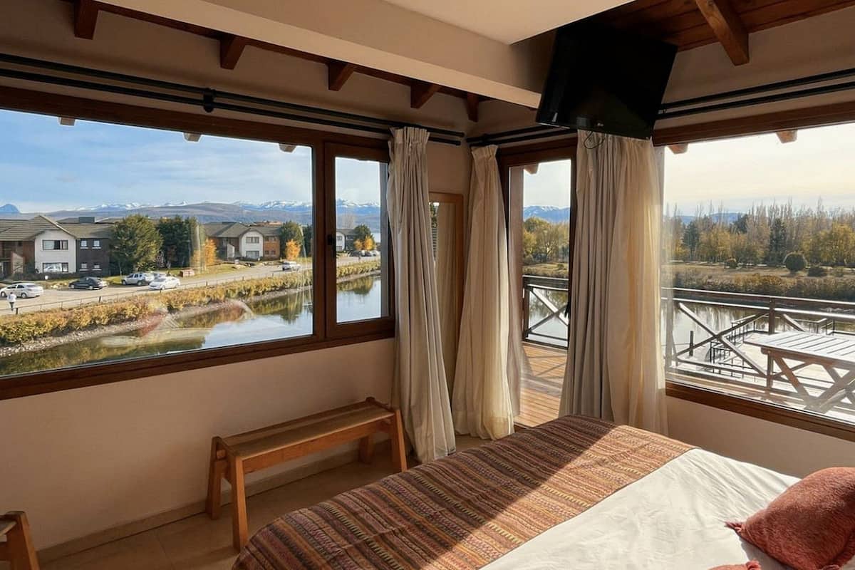 Un dormitorio con una cama grande, vigas de madera y vistas a un lago y edificios.