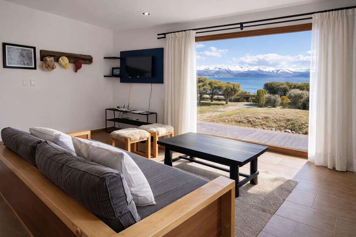 Una sala moderna con sofá, una mesita de centro y televisor montado en la pared. Hay un ventanal con vistas a montañas y una masa de agua.