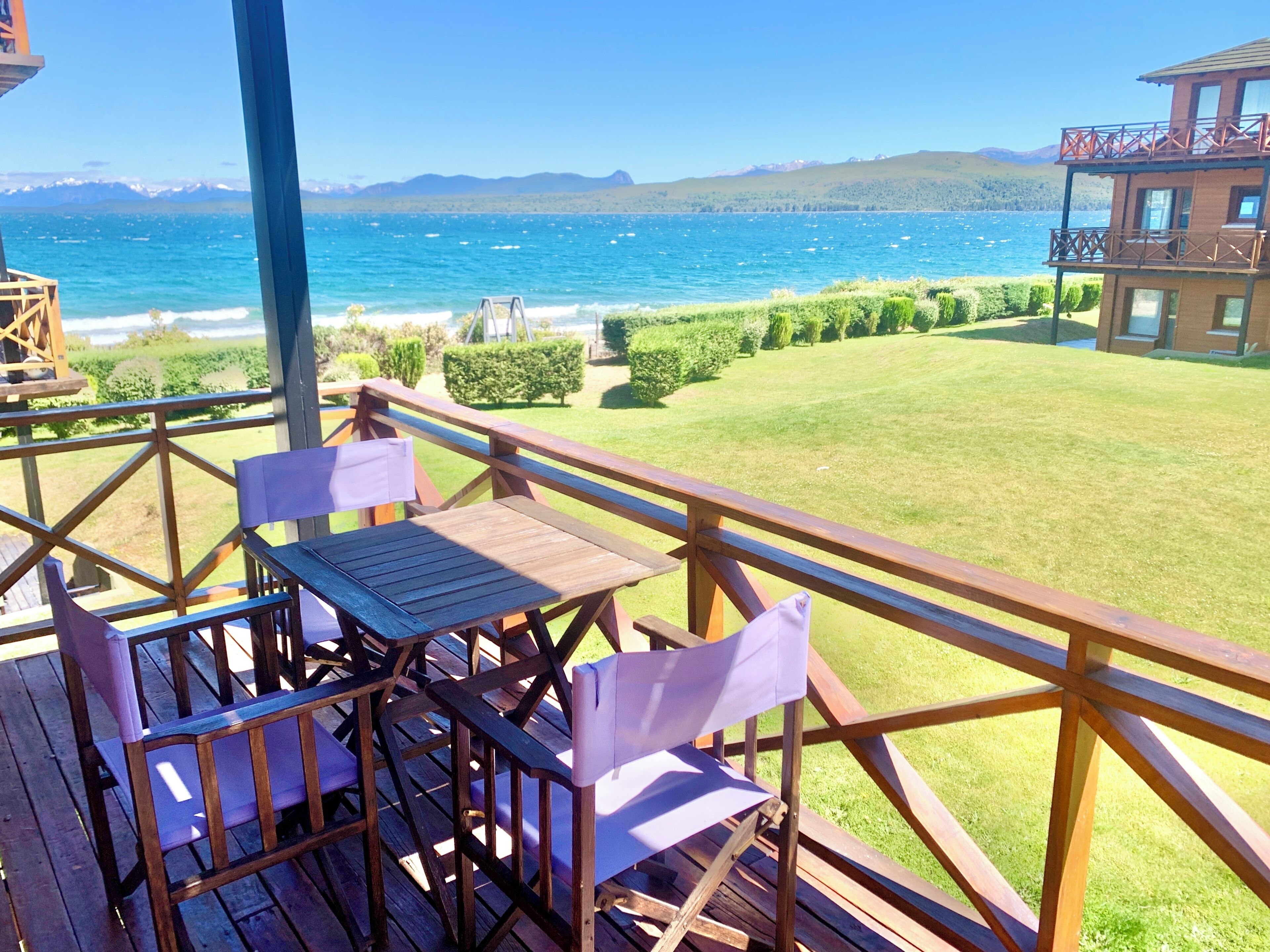 Una terraza de madera con una mesa y sillas que da a un lago con barcos, rodeada de follaje y montañas a lo lejos.
