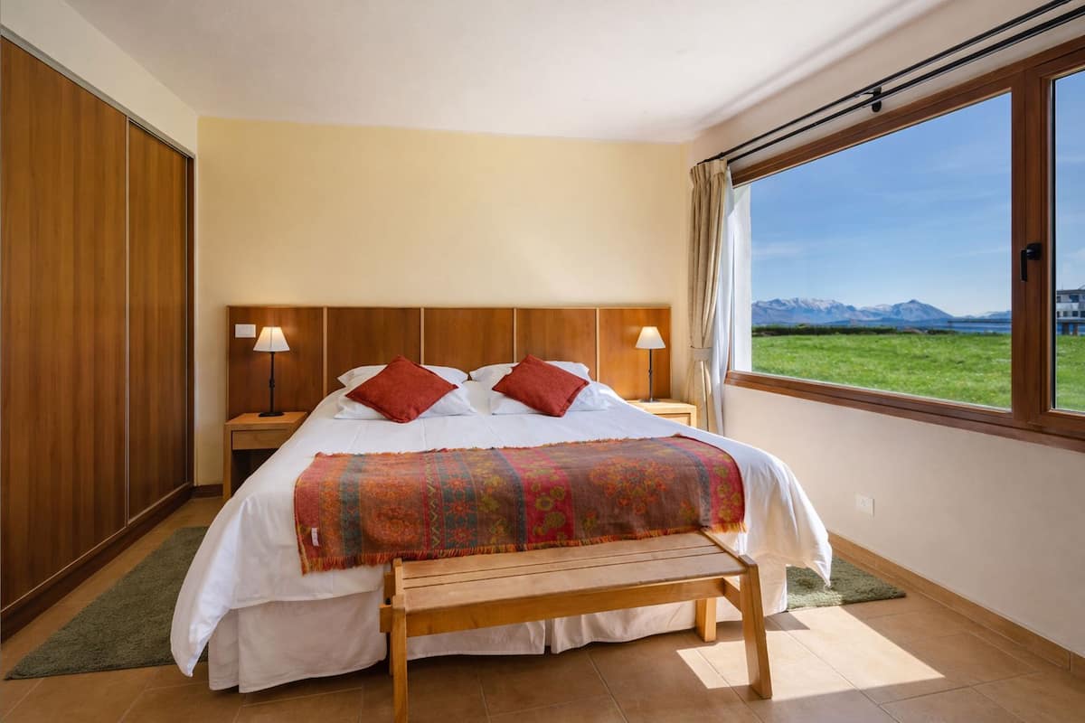 Un dormitorio con una cama grande, dos lámparas de noche, un banco, vistas a montañas y un armario de madera.