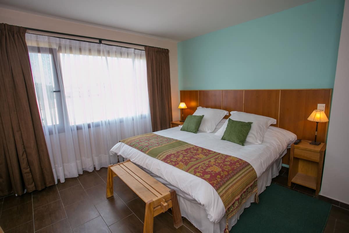 Una habitación de hotel con una cama grande, un banco de madera, una mesita de noche con lámpara y una ventana con cortinas.