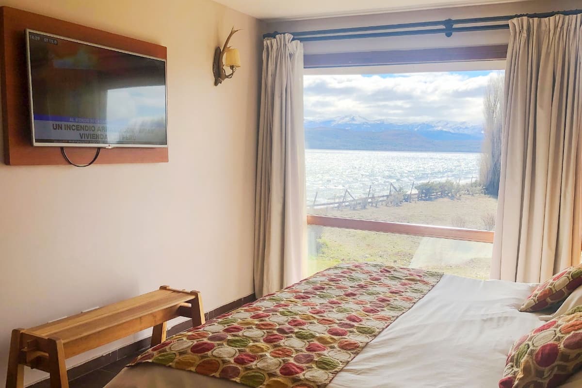 Un dormitorio con cama, una banca, televisor montado y vistas a un lago y montañas.