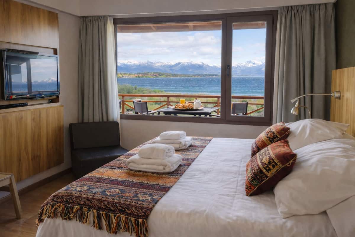 Habitación de hotel con cama, televisión, mesa con desayuno y vistas a montañas y agua.