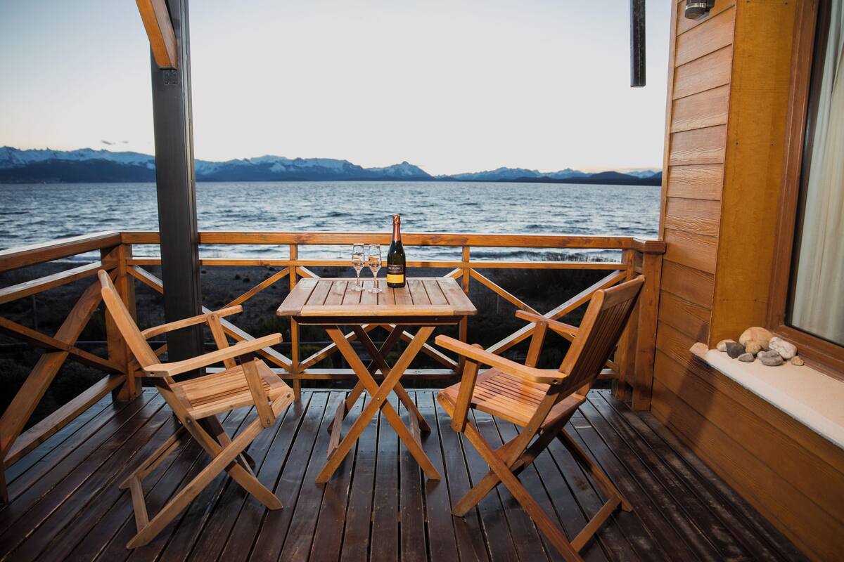 Una terraza de madera con una mesa y sillas, con vista a un cuerpo de agua y montañas al fondo.