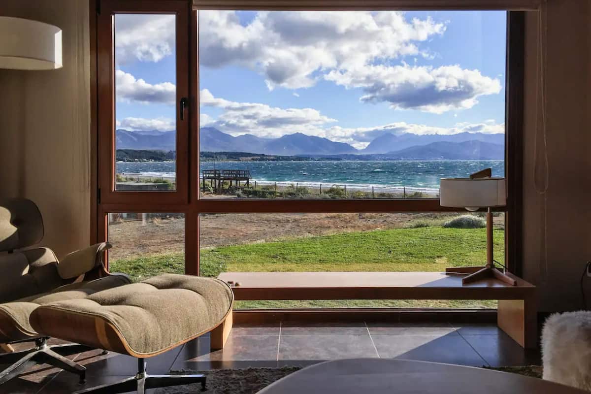 Habitación con un ventanal grande que ofrece vistas al mar y a las montañas. Hay una silla y una lámpara en el suelo.