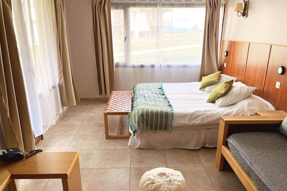 Una habitación de hotel con cama, mesita de noche, banco y una pequeña alfombra blanca y esponjosa.