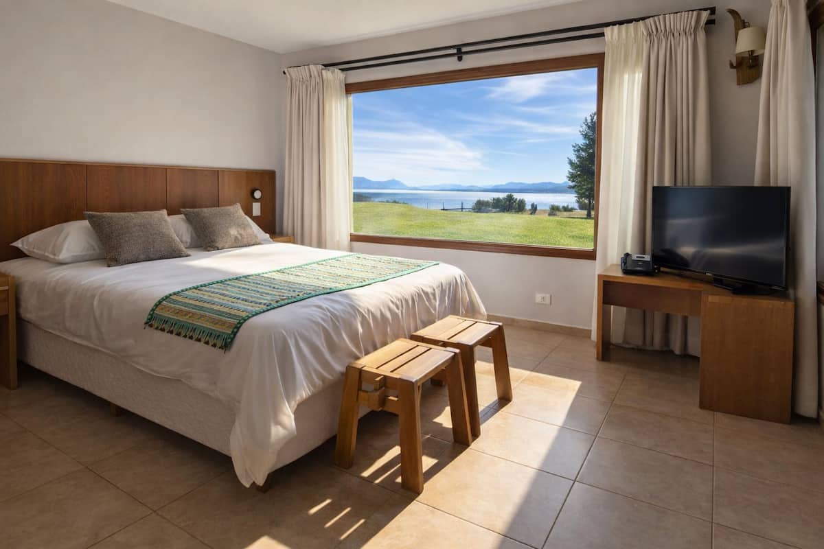Un dormitorio con una cama, mesitas de noche de madera, un televisor, una ventana con vista a un lago y montañas, y un taburete pequeño.