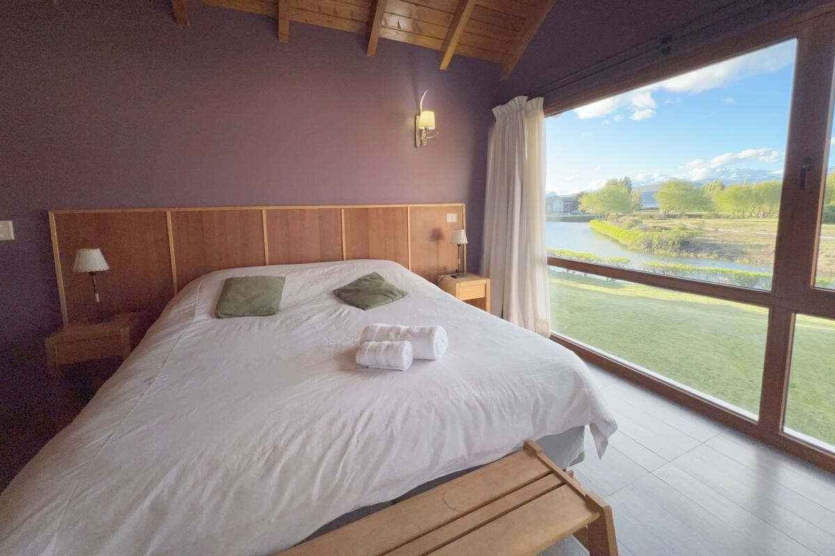 Un dormitorio con una cama grande, cabecera de madera, dos lámparas de noche, una mesita de noche y vistas a un lago y vegetación.