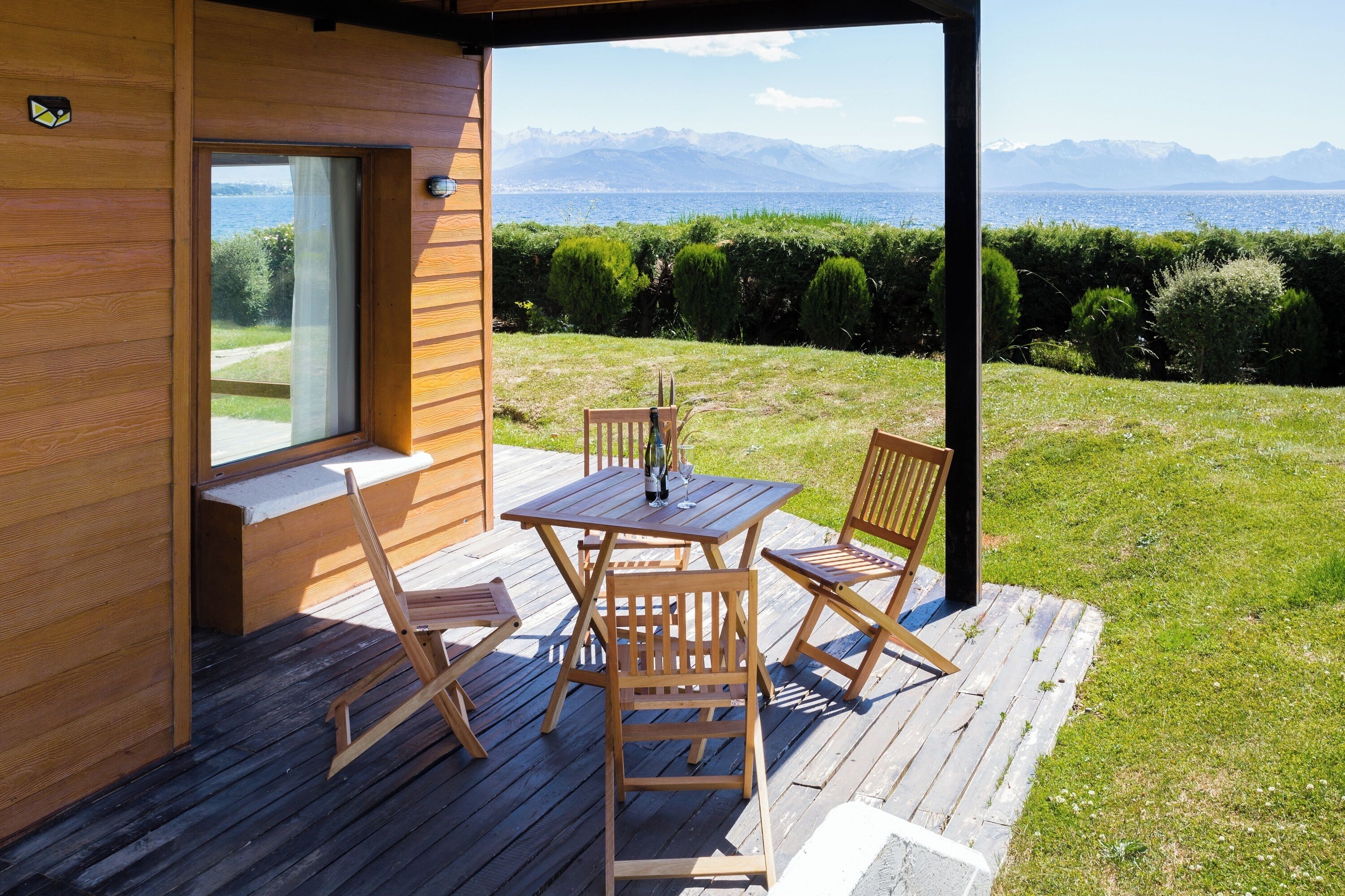 A wooden deck with a table and chairs, a window, and a view of mountains and water.