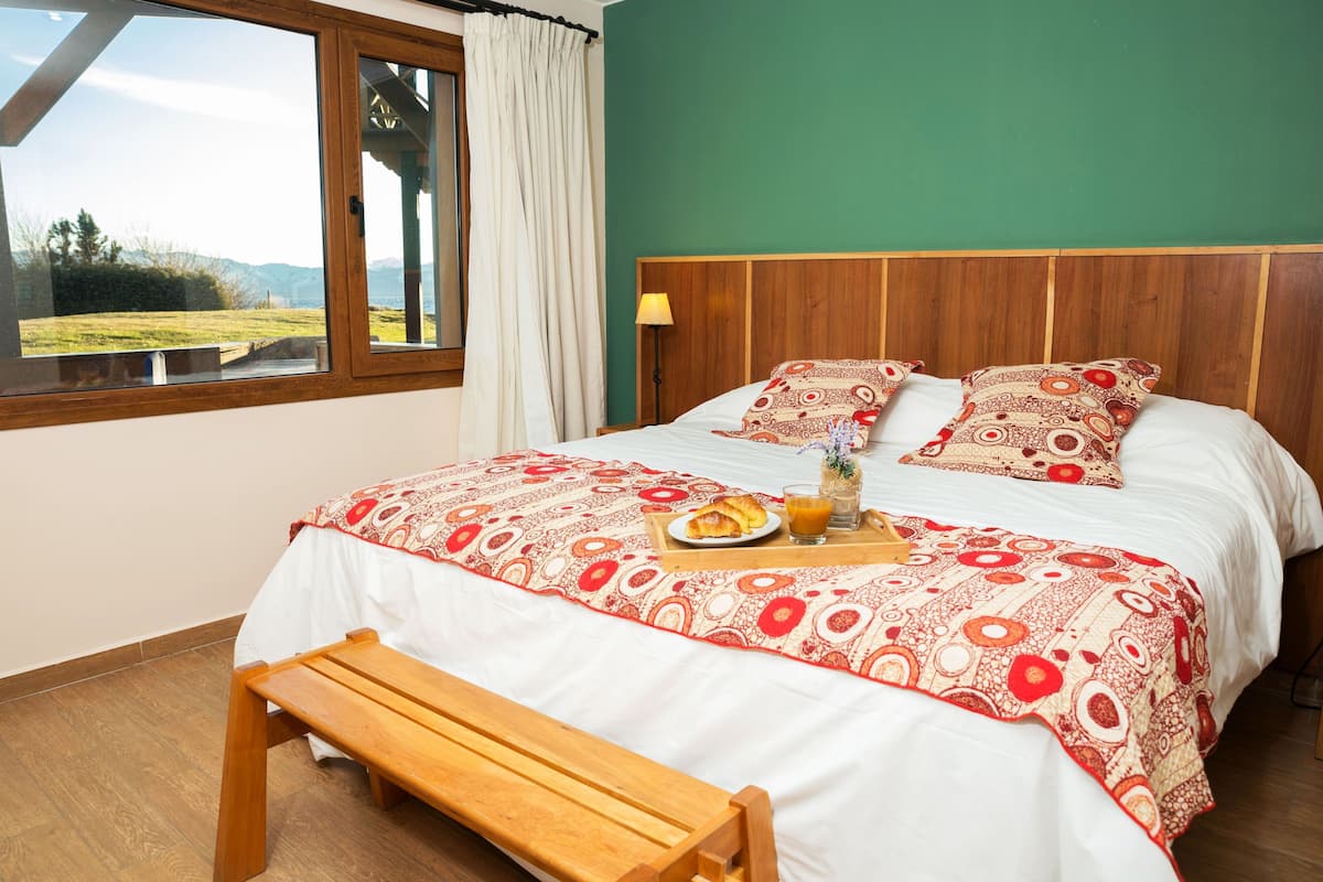 Un dormitorio con cama y banco de madera, una bandeja de desayuno con tostadas y jugo, y vistas a un paisaje verde a través de una ventana.