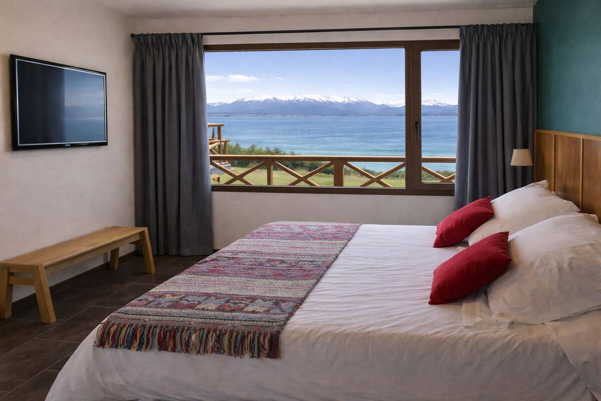 Un dormitorio con una cama grande, televisor, una banca y vistas a un lago y montañas.